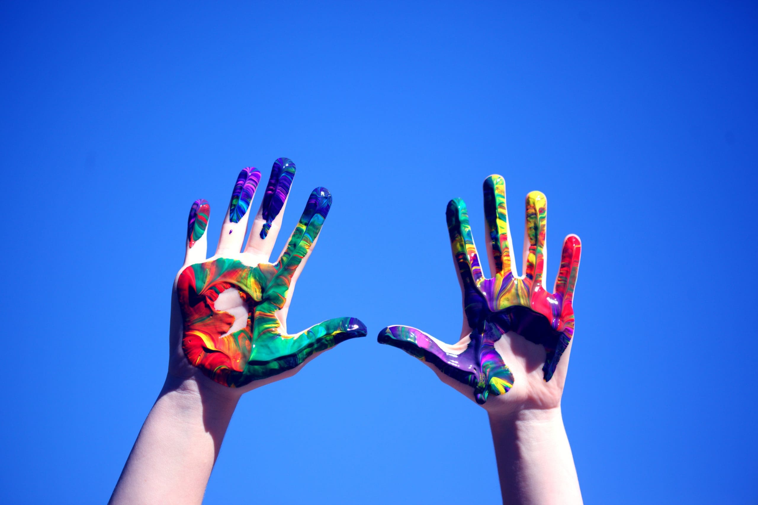 Two hands painted with colors are raised against a blue sky.