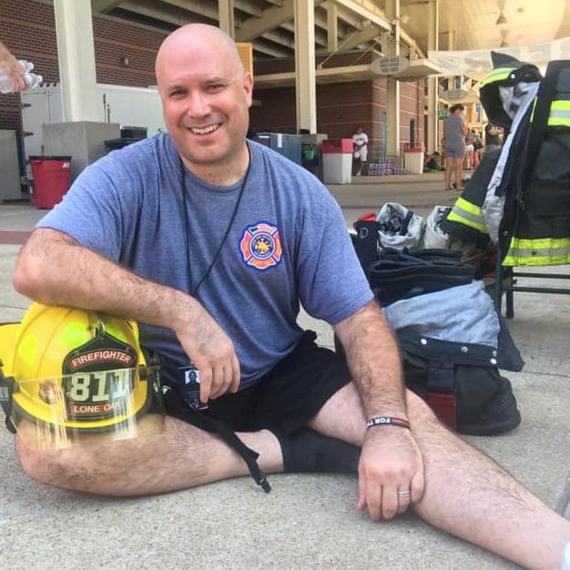 John smiles and sits on the pavement with his firefighting gear at his side.